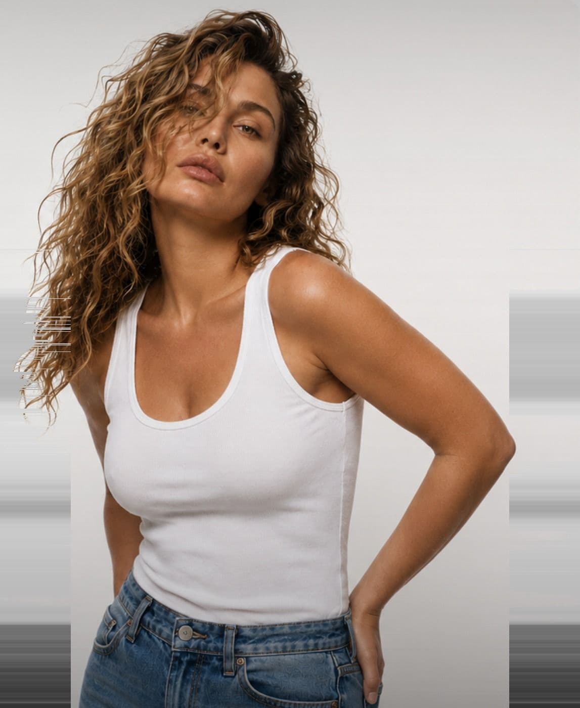 A woman in a white tank top and jeans, hands on her hips, hair loose